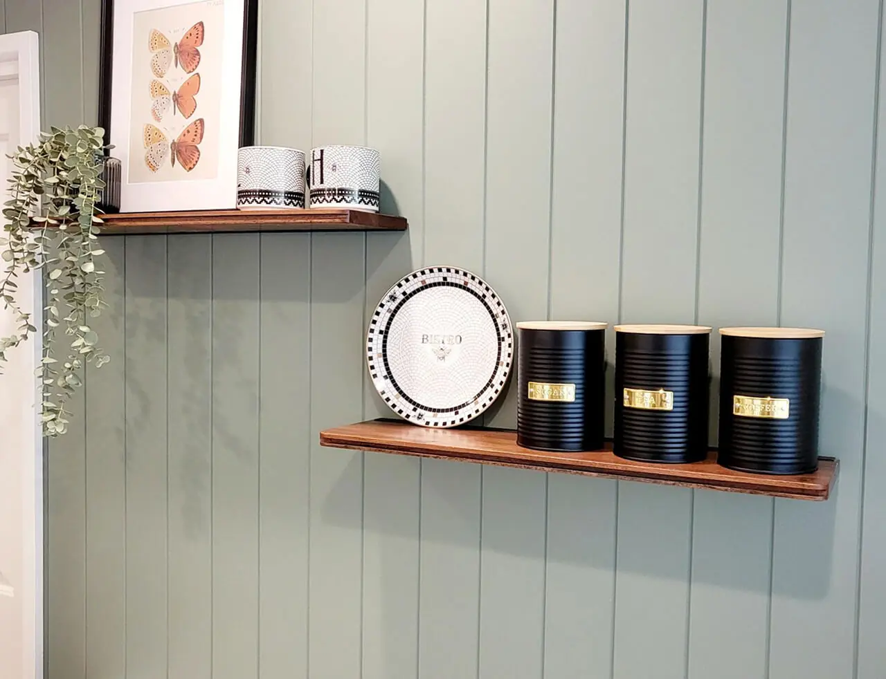 odern kitchen shelving decor with black canisters, a decorative bee plate, and trailing greenery on a sage green paneled wall.
